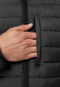 Jachetă puf cu model capitonat, de culoare neagră, dotată cu un buzunar lateral cu fermoar. O mână întinsă în buzunar, evidențiind textura netedă.