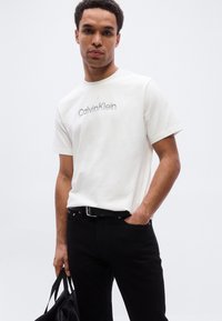 Λευκό βαμβακερό T-shirt με το λογότυπο Calvin Klein σε γκρι, συνδυασμένο με μαύρο τζιν και μαύρη τσάντα, στέκεται σε ουδέτερο φόντο.
