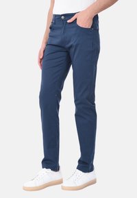 Pantaloni in cotone blu navy con una vestibilità slim, dotati di chiusura con bottone, tasche laterali e una texture liscia. Abbinati a sneakers bianche.