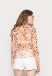 Camisola de mangas longas em tecido transparente com um padrão em espiral nas cores laranja, bege e branco. Apresenta um laço na cintura e mangas largas, combinada com calções brancos.