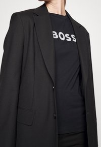 Blazer negro con un diseño estructurado y solapas de muesca, superpuesto a una camiseta negra con la palabra "BOSS" en letras blancas y en negrita.