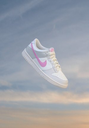 Baskets Nike blanches et grises avec un swoosh et des lacets roses sur un fond de ciel doux bleu et orange.