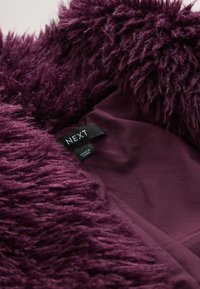 Lila Kunstpelzstoff mit langen, weichen Fasern, der eine glatte Innenfläche zeigt. Das Etikett trägt die Aufschrift "NEXT" und verweist auf Marke und Herkunft.