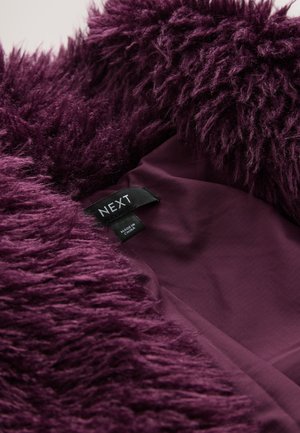 Violets mākslīgās kažokādas audums ar gariem, mīkstiem diegiem, atklājot gludu iekšējo oderi. Etiķete satur uzrakstu "NEXT", norādot zīmolu un izcelsmi.