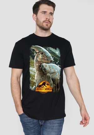 Mann trägt ein schwarzes Jurassic World T-Shirt mit einem Parasaurolophus-Dinosaurier und Logo, steht mit einer Hand in der Tasche.