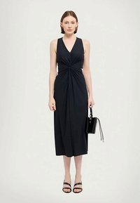 Ermeløs, marineblå kjole med vridd detalj i midjen, kne-lengde, laget av glatt stoff, kombinert med en liten svart håndveske og sandaler.