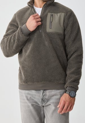 Homme portant un pull polaire gris avec une poche poitrine zippée et un jean gris clair, ajustant le col avec la main gauche visible portant une montre noire.