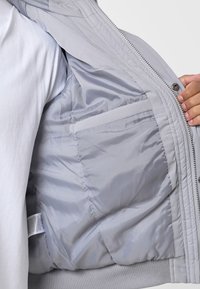 Svetlošedá puffová bunda s prešívaným vnútrom, rebrovaným lemom a bočnou kapsou. Vyniká hladkým exteriérom so zipsovým uzáverom.