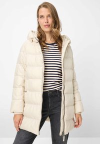 Lyse beige dunjakke med hætte, der har lynlås foran og lodret syningsmønster. Bærres over en navy- og hvidstribet skjorte og mørke jeans.