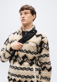 Pull polaire beige et noir à motif, avec un col montant et une poche avant, présentant une bande contrastante noire sur l'épaule.