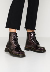 Botas de tobillo de cordones en charol burdeos brillante con cordones negros, que presentan una suela cosida en amarillo y una suela de diseño rugoso.