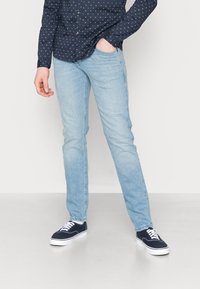 Man som bär ljusblå slim-fit jeans, marinblå sneakers med vita snören, och en mörkblå långärmad skjorta med ett litet vitt mönster.