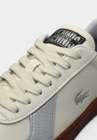 Αθλητικό παπούτσι από λευκό δέρμα με υφασμένο ασημένιο πλαϊνό μέρος, κρεμ κορδόνια και μαύρη ετικέτα με λογότυπο. Διαθέτει λάστιχο σόλα με καφέ λεπτομέρειες.