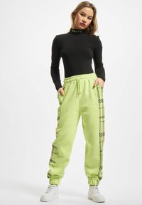 Svart langermet topp med teksten "Thug Life"; neongrønne joggebukser med svart geometrisk sidedesign, elastisk liv og mansjetter. Hvite sneakers.