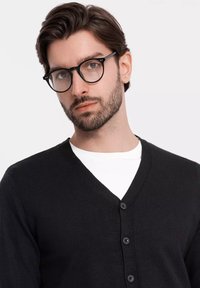 Svart cardigan med V-ringad design, med fem svarta knappar. Bärs över en vit t-shirt, med en slät och mjuk textur.
