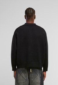 Schwarzer Strickpullover mit weitem Ausschnitt, überschnittenen Schultern und gerippten Bündchen. Kombiniert mit grauen, locker sitzenden Jeans.