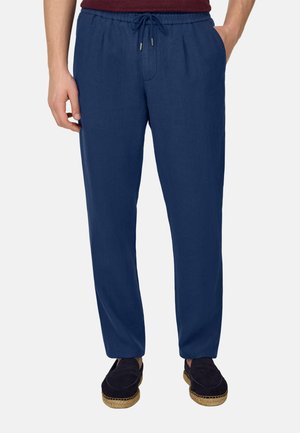 Mand iført marineblå snøre bukser og mørke slip-on sko, stående med hænderne i lommerne mod en ensfarvet baggrund.