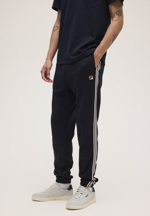 Uomo che indossa pantaloni della tuta neri con strisce laterali bianche e rosse, scarpe da ginnastica bianche e una maglietta nera, in piedi con una mano in tasca.