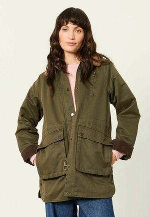 Frau mit langen dunklen Haaren, die eine olivgrüne Jacke mit großen Taschen und braunen Bündchen trägt, steht mit den Händen in den Taschen vor einem einfarbigen Hintergrund.