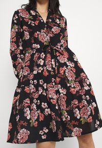 Květinové šaty z černé tkaniny s velkými růžovými a červenými květinami, s přiléhavým korzetem, zavázaným pasem a rozšířenou sukní s vrstvenými volány.