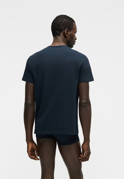 Moški, ki stoji z obračnim hrbtom, oblečen v temno modro majico in ujemajoče prilegajoče se kratke hlače, na belem ozadju.