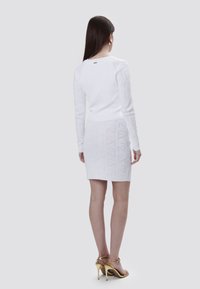 Witte gebreide top met lange mouwen en textuurpatroon, gecombineerd met een witte minirok met een geometrisch ontwerp, aangevuld met gele haksandalen.