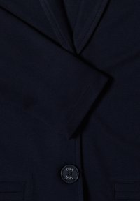 Gros plan de la manche d'un blazer bleu marine foncé, du revers et d'un seul bouton noir portant l'étiquette "Cecil."