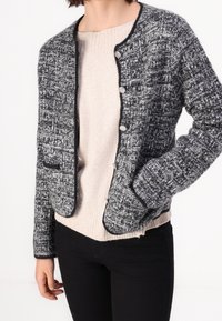 Giacca grigia e nera con texture, scollo rotondo, cinque bottoni d'argento e piping nero, indossata sopra un maglione color crema.