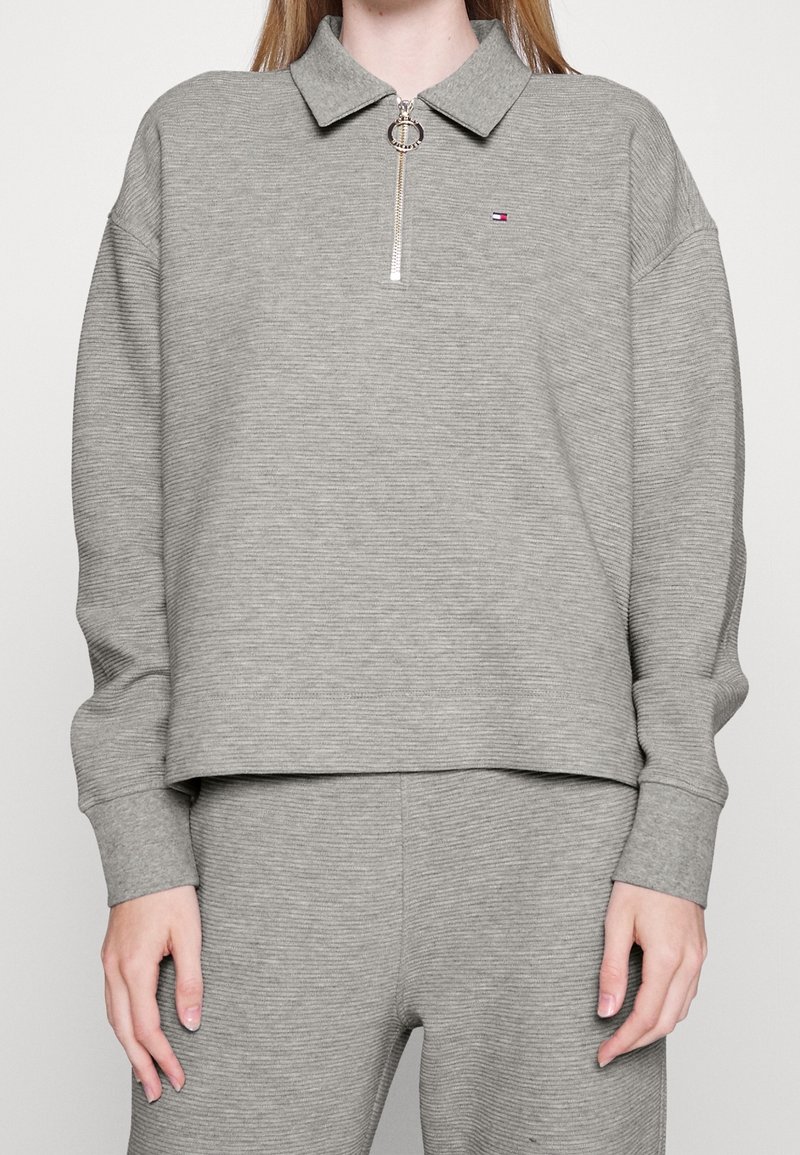 Femme portant un pull à manches longues en maille côtelée gris avec une fermeture éclair à moitié et un petit logo sur la poitrine gauche, associé à un pantalon côtelé assorti.