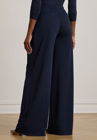 Asmuo vilkintis tamsiai mėlynas plačias kelnes ir atitinkamą ilgą rankovę, stovintis ant medinių grindų prieš paprastą sieną.