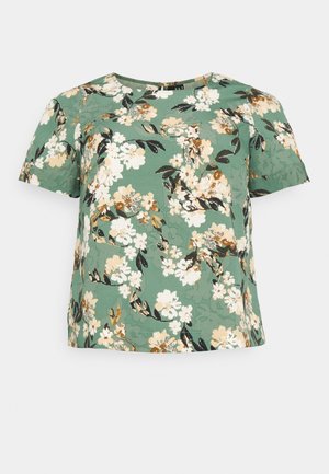 Blouse verte à manches courtes avec col rond, présentant un motif floral avec des fleurs blanches, beiges et noires ainsi que des feuilles.