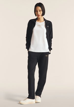 Giacca nera con zip sopra una t-shirt bianca, abbinata a pantaloni tuta neri e sneakers bianche. Presenta scritte dorate sulla maglietta e sui pantaloni.
