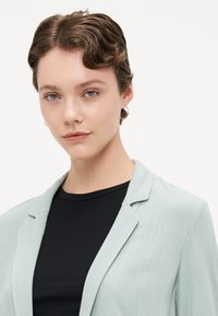 Hellgrüner Blazer mit Revers und strukturierter Oberfläche, getragen über einem schwarzen rippenförmigen Oberteil. Das Model hat kurzes, gewelltes Haar und Ohrringe mit Creolen.
