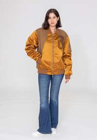 Braune und goldene Satin-Bomberjacke mit einem Frontreißverschluss, gerippten Bündchen und besticktem "8"-Design. Getragen über Jeans im Schlag.