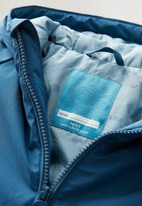 Blauwe jassenbinnenkant met een ritsluiting, zachte voering en een label met de tekst "NEXT" met zorginformatie en een decoratief logo.