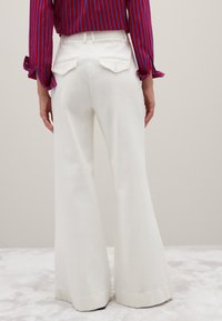 Donna che indossa pantaloni bianchi a gamba larga con tasche posteriori abbottonate e una camicia a maniche lunghe a strisce rosse e blu, in piedi su un tappeto chiaro.