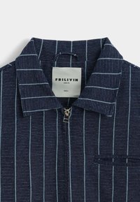 Chemise bleu marine à fermeture éclair avec des rayures verticales blanches, un col pointu et une petite poche poitrine. L'étiquette indique "FRILIVIN, PARIS, FR, TAILLE L."