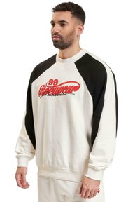 Hvit sweatshirt med svarte raglanermer, med rød broderi tekst "99 Babewear" på brystet. Myk, avslappet passform, rund hals.