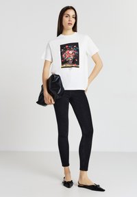 Donna che indossa una t-shirt bianca con grafica floreale, jeans skinny neri, scarpe basse a punta nere, mentre tiene una borsa a mano nera, in piedi di fronte a uno sfondo uniforme.