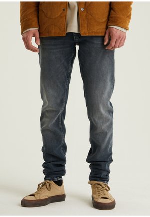 Jeans de mezclilla azul oscuro con un ajuste slim, ligeros desgastes y diseño de cinco bolsillos; combinados con zapatillas de gamuza claras y una chaqueta de pana.