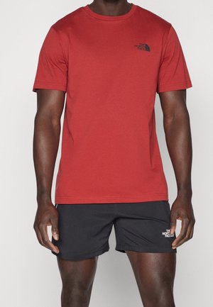 Ein Mann trägt ein rotes T-Shirt mit kurzen Ärmeln und schwarze Shorts, beide mit dem Logo von The North Face auf der Brust bzw. dem Bein.