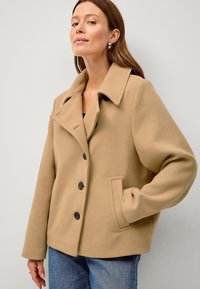 Beige uld-blandingsjakke med spidse klapper, sorte knapper og sidelommer. Glat tekstur og afslappet pasform, parret med denim.