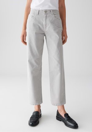 Vrouw draagt lichtgrijze straight-leg jeans en zwarte leren loafers, staand tegen een effen witte achtergrond.