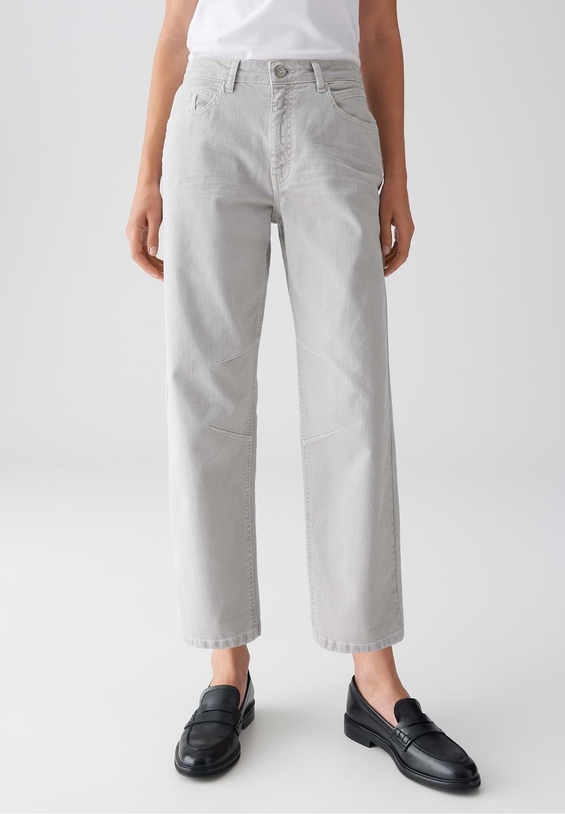 Femme portant un jean droit gris clair et des mocassins en cuir noir, debout devant un fond blanc uni.