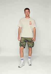 Jovem em pé, vestindo uma t-shirt gráfica beje, calças cargo com padrão de camuflagem, meias brancas com riscas vermelhas e azuis, e sapatilhas brancas.