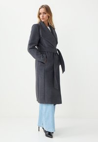 Grijze lange jas met een wikkelontwerp, brede revers, strik taille en zijzakken. Gemaakt van een zachte, gestructureerde stof. Gecombineerd met lichtblauwe wijde jeans.