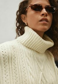 Cremfarbener Kabelstrickpullover mit geripptem Rollkragen. Das Modell trägt braune Cat-Eye-Sonnenbrille und hat lockiges Haar. Strukturierte Stoffdetails sichtbar.