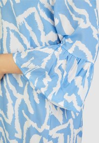 Hellblaue und weiße Bluse mit Muster, die aus einem strukturierten Stoff gefertigt ist. Sie hat weit ausgestellte Ärmel und ein abstraktes Design mit unregelmäßigen Linien und Formen.