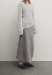 Personne portant un pull en maille gris clair, une longue jupe grise portefeuille, des chaussettes beiges et des mocassins noirs, debout contre un mur uni.