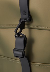 Détail d'un sac imperméable vert olive avec une boucle en plastique noir et une sangle, des accents cousus, et une texture lisse et mate.
